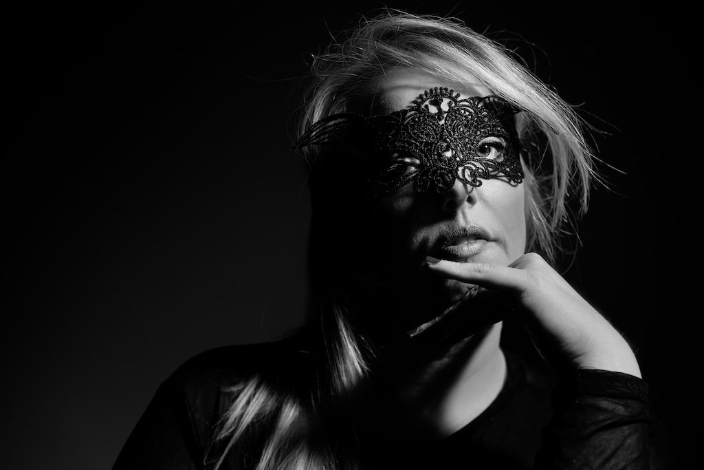 Black and white portrait of a blonde woman wearing an ornate masquerade mask, posing dramatically.