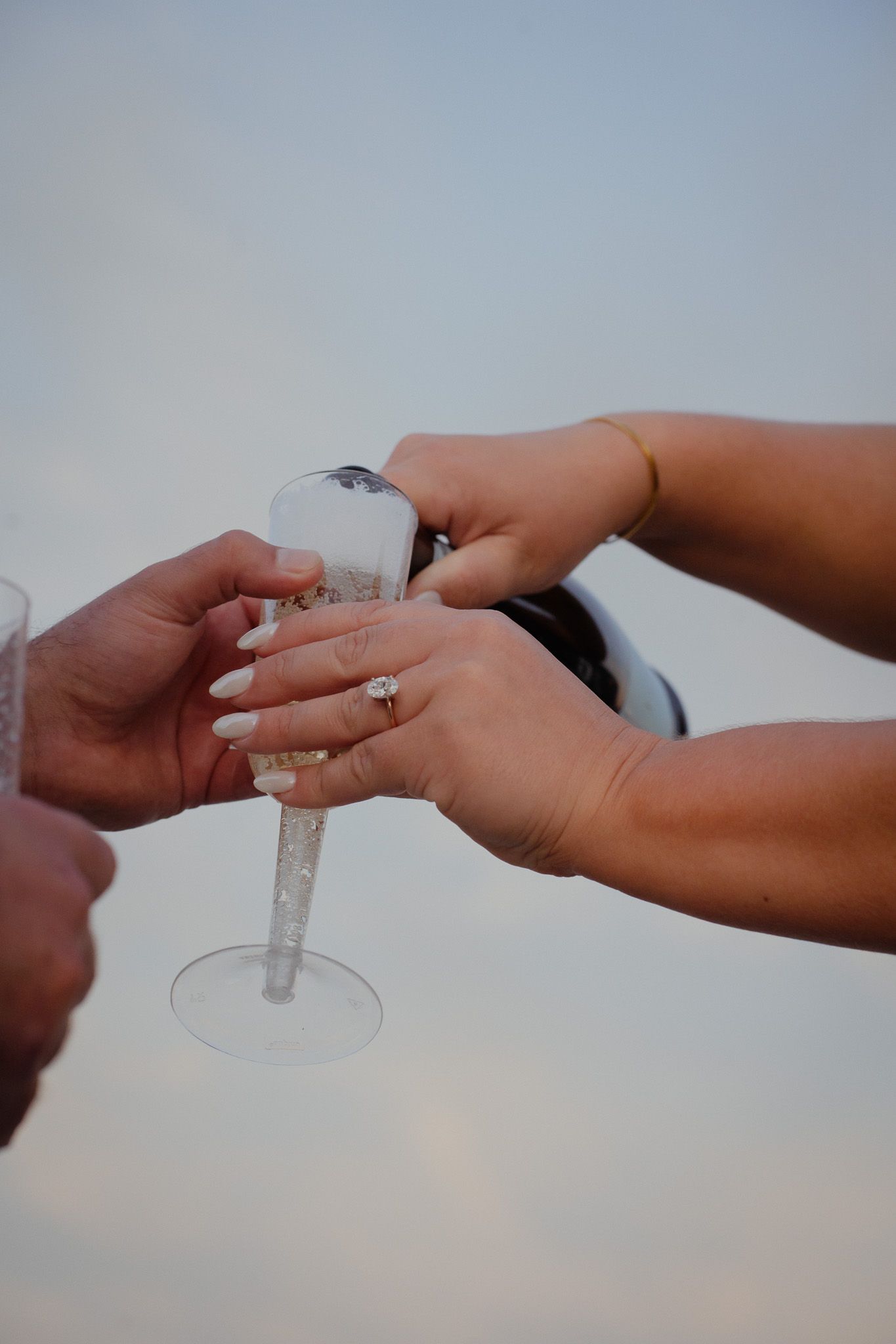 Two pairs of hands holding a champagne glass and filling it up with champagne. Ring is on the soon to be brides hand