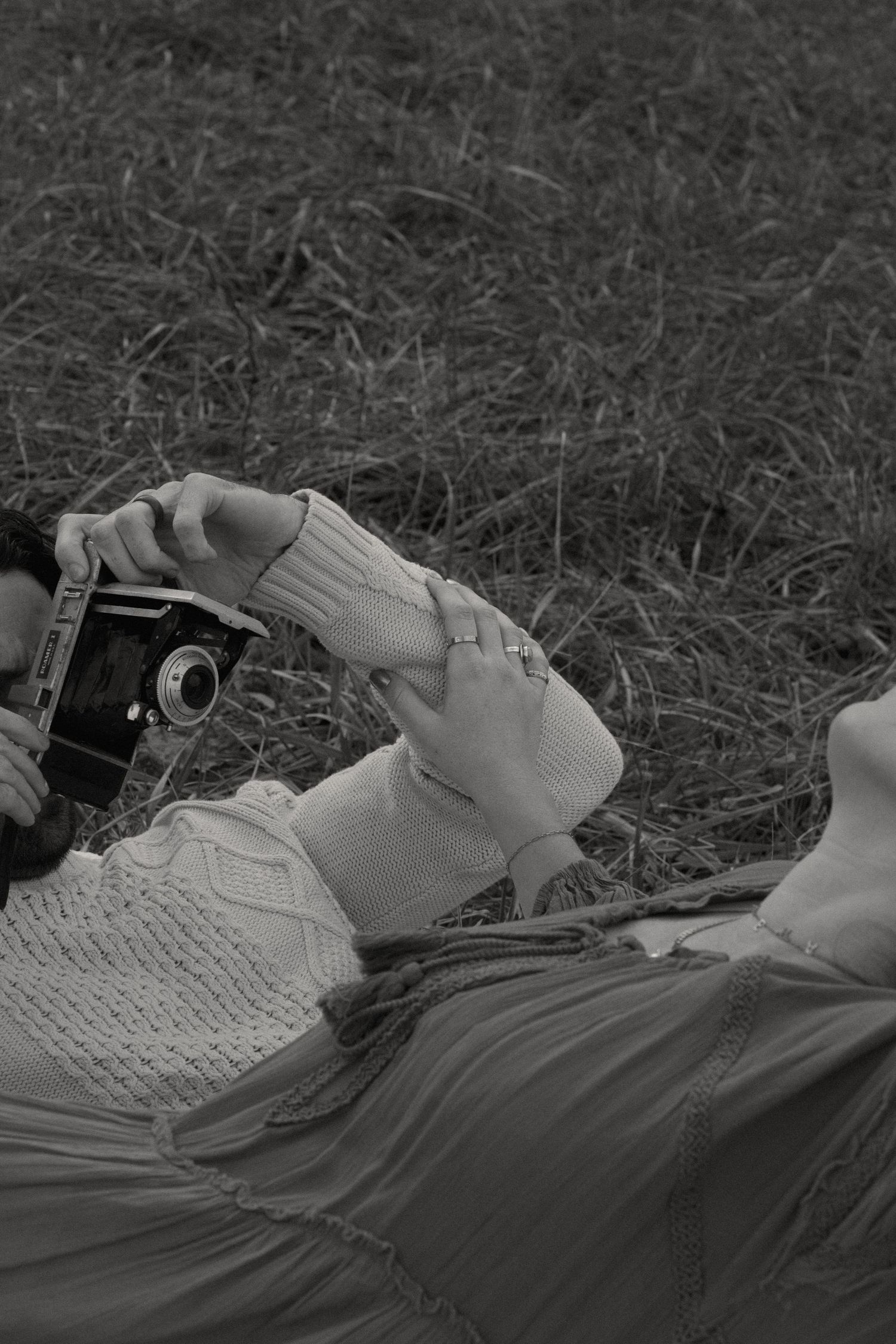 Woman laying on the grass with her head tilted back. Her hand is on her husbands arm while he is trying to take her pict