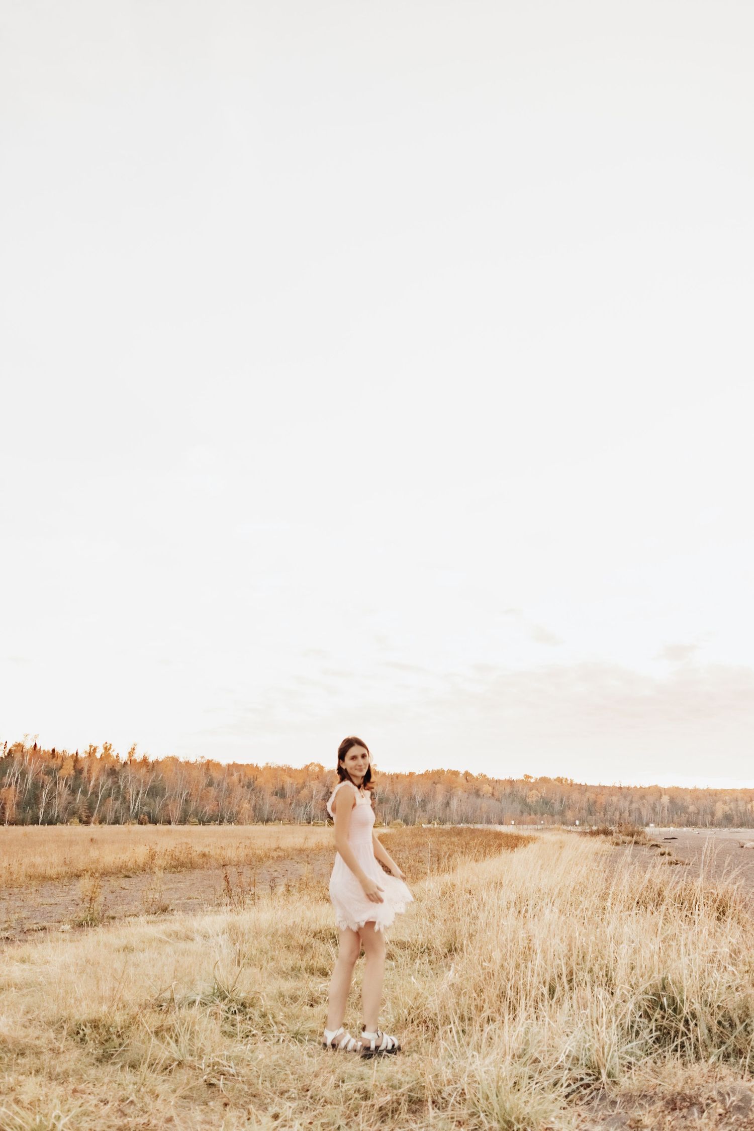 Young woman in white dress standing in golden autumn field with trees in background.