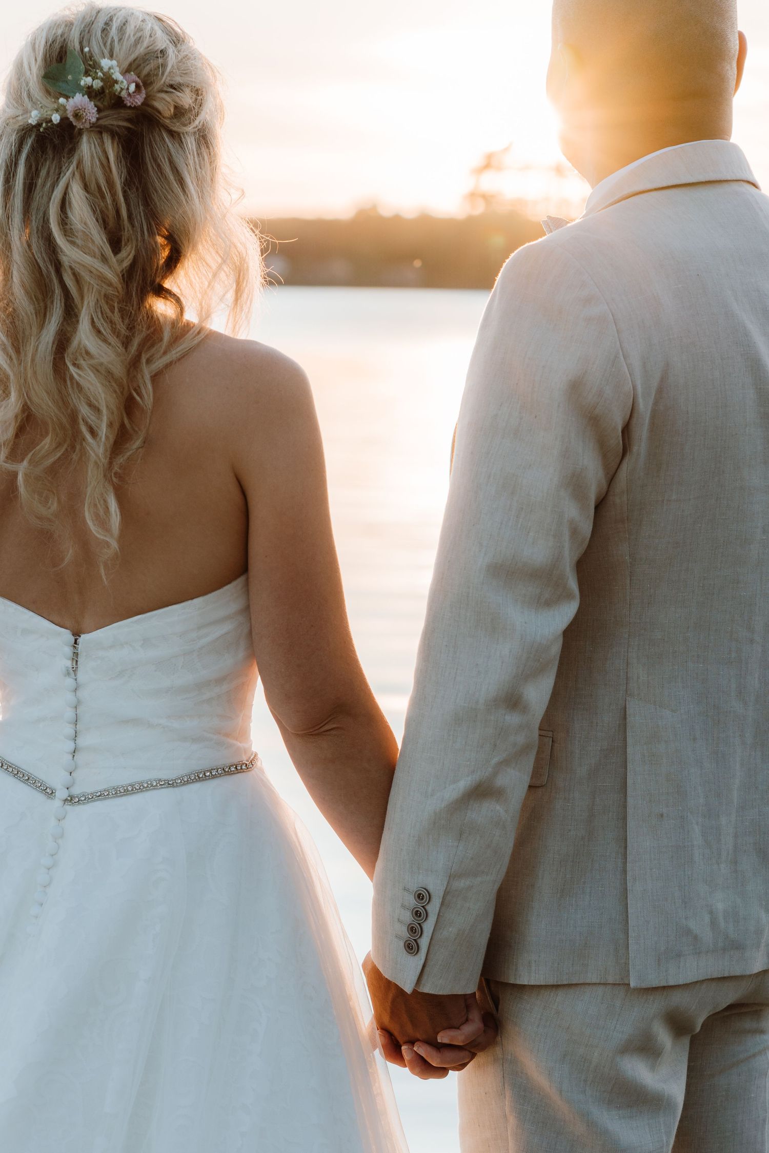 A couple in wedding attire stands together at sunset, with warm golden light illuminating their silhouettes.