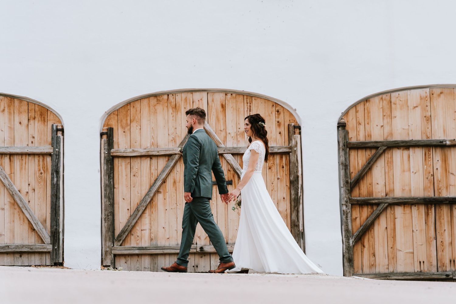 A couple in wedding attire walking past rustic wooden barn doors against a white wall.