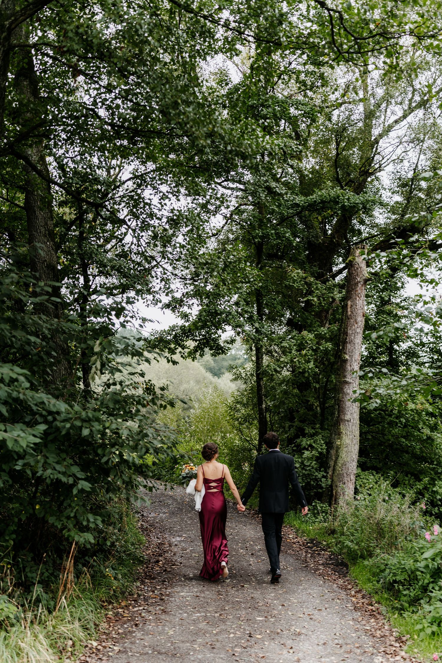 Bildaussage: Der Hochzeitstag gehört dem Brautpaar. Dieses läuft Hand in Hand, entfernt durch eine grüne, bewaldete Szenerische Landschaft.
