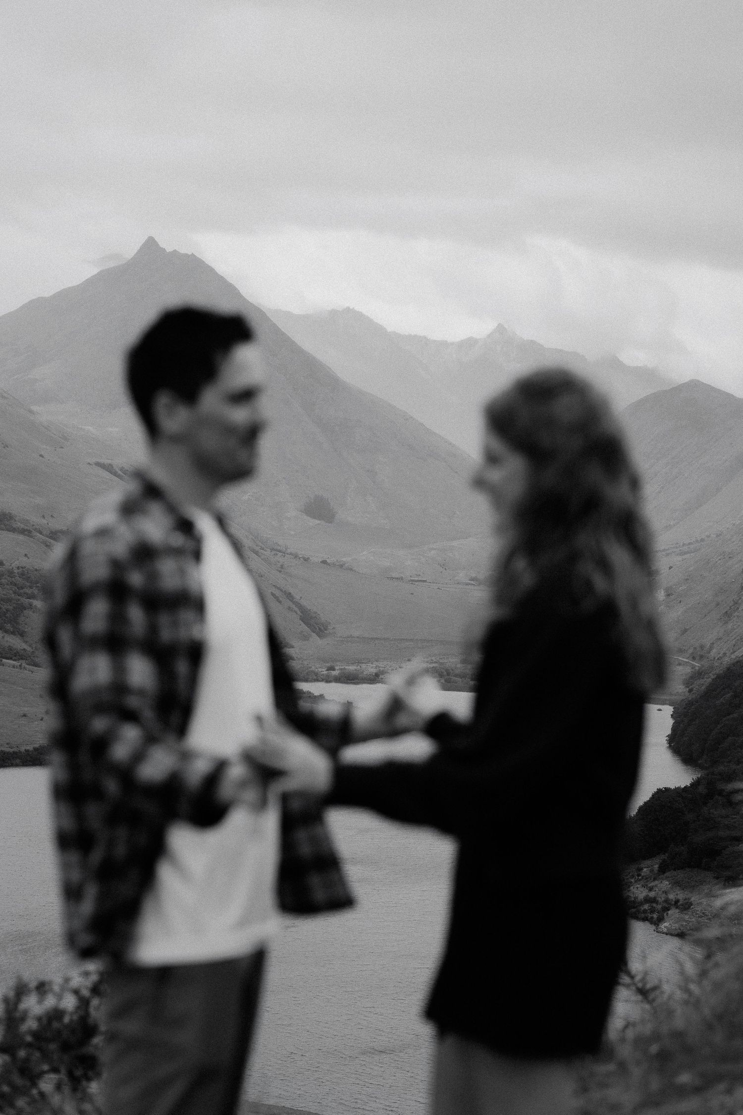 Black and white photo of a couple holding hands with mountains and a lake in the background.