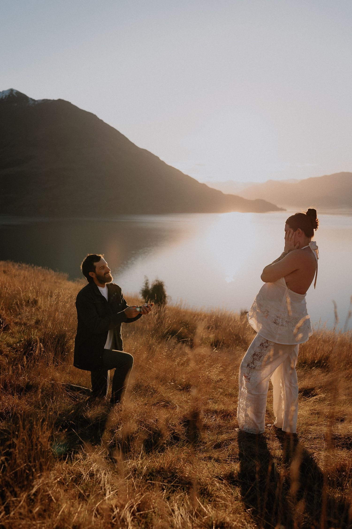 Man kneeling to propose to woman by a mountain lake at golden hour sunset in a scenic outdoor setting.