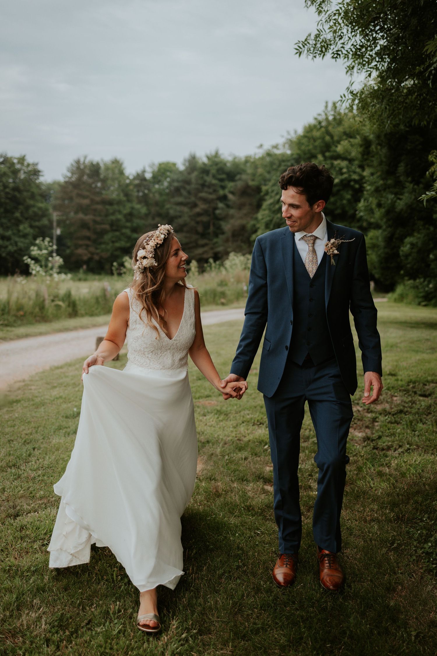 Newlywed couple walking and laughing together on grassy path with forest backdrop.