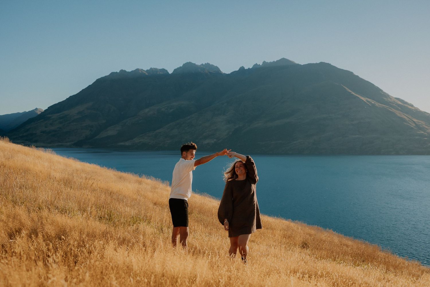 Couple dancing on golden hillside with stunning mountain lake backdrop under clear blue sky.