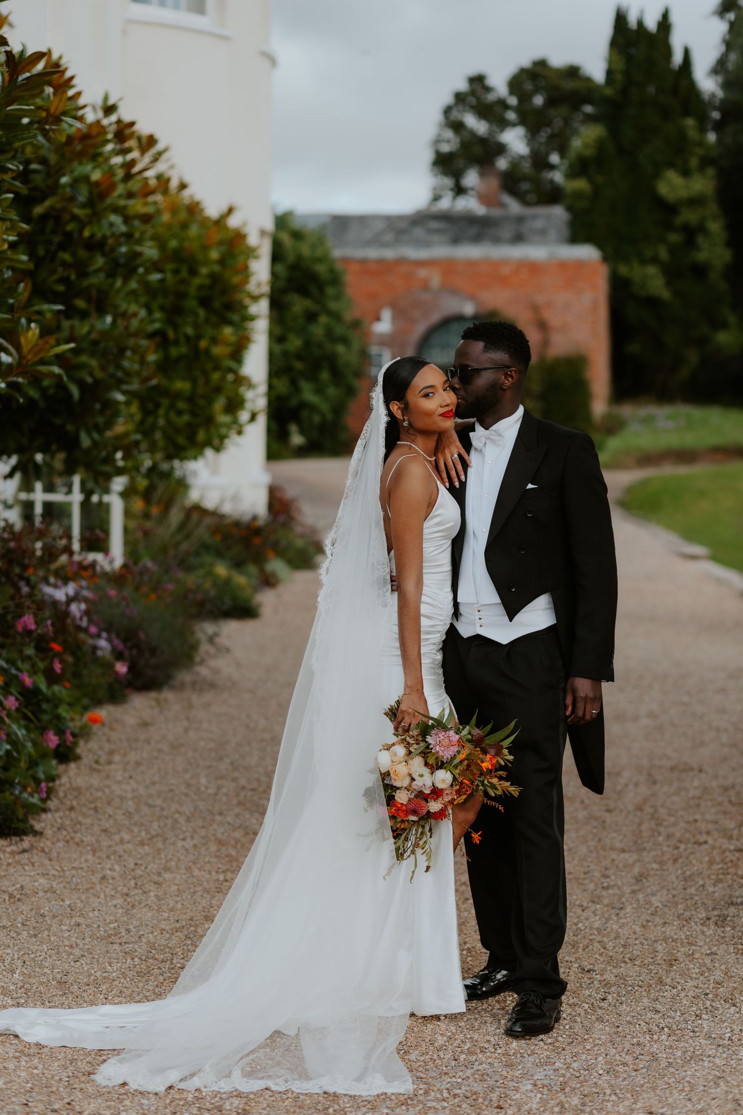 Elegant bride and groom embrace on garden path while holding stunning floral bouquet.