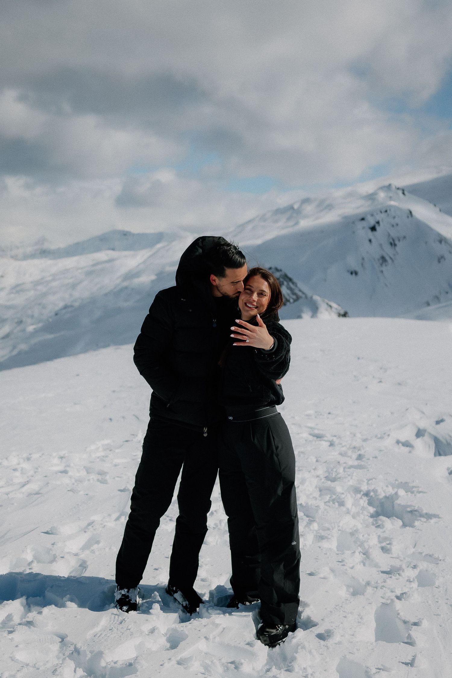 Couple embracing on snowy mountain summit, wearing black winter outfits, with scenic snow-capped peaks and cloudy sky behind them.