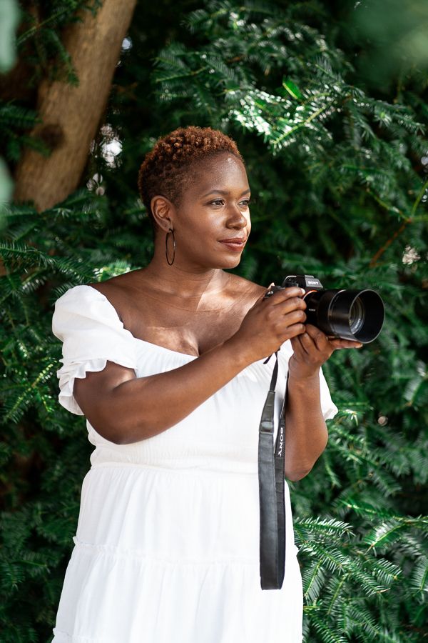 A photographer in a white dress holding a camera in a lush garden setting.