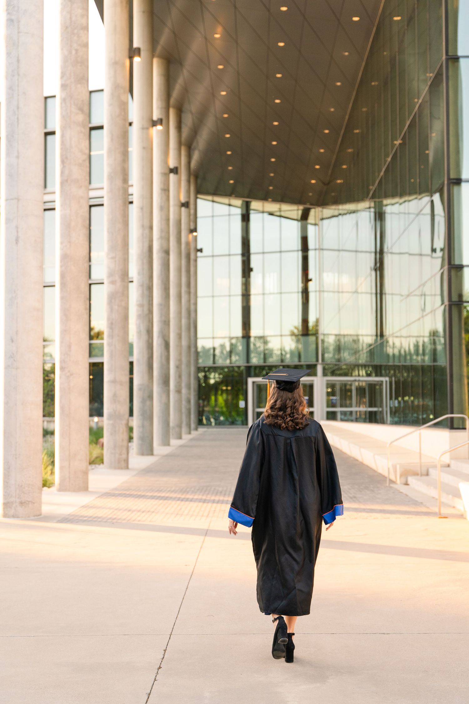 Grad Session in DFW, Texas. Girl in a black cap and gown walks away down a walkway with white columns at UTA Arlington.