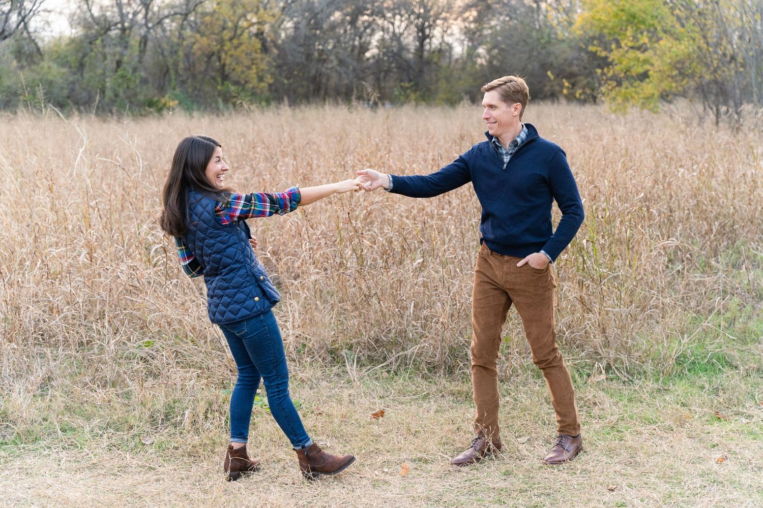 Couple Session in DFW, Texas. Wife holds her husband's hand and leans back as they smile and dance together in a field of tall dry grass.