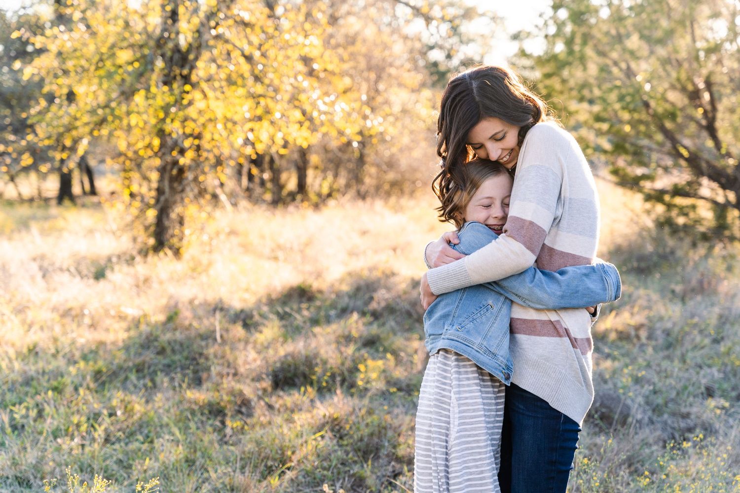 Family Session in DFW, Texas. Mom and her young daughter hug each other as sunlight streams through the fall leaves behind them.