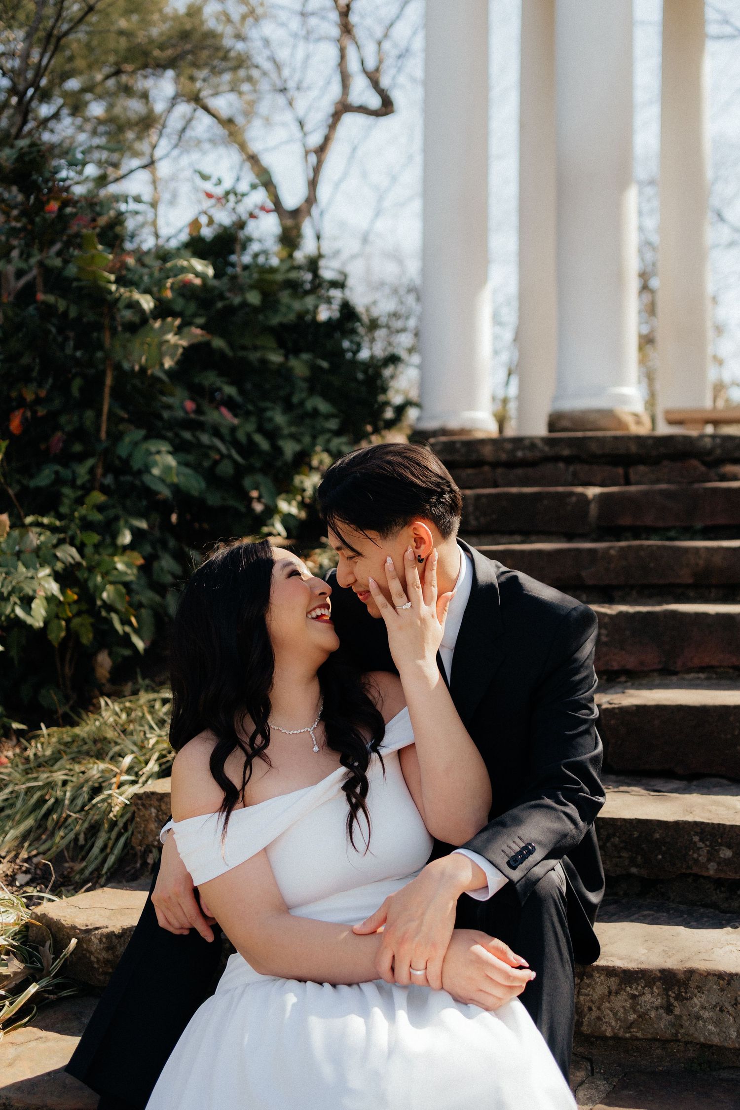 Bride and groom sitting on stone steps, sharing a romantic moment near white columns and lush greenery.