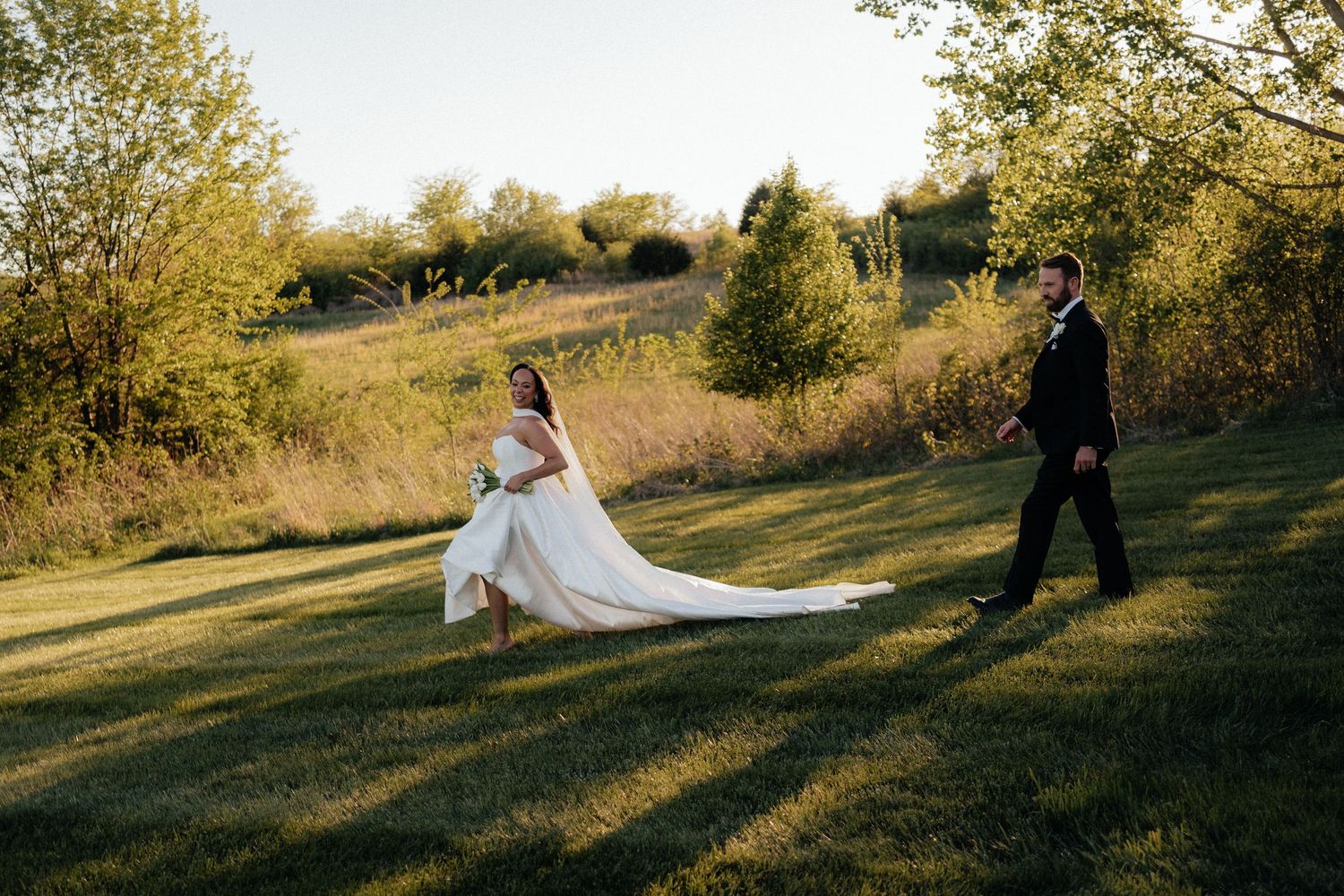 Bride and groom walk across lush green lawn at golden hour, wedding dress train flowing behind them.