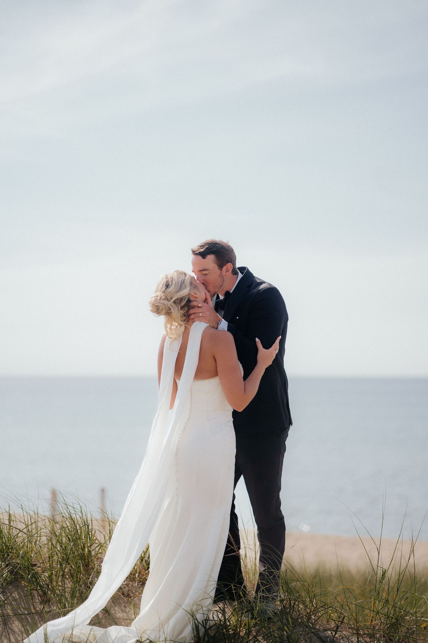 A romantic wedding couple shares a tender kiss atop a grassy coastal cliff overlooking the ocean on their special day.