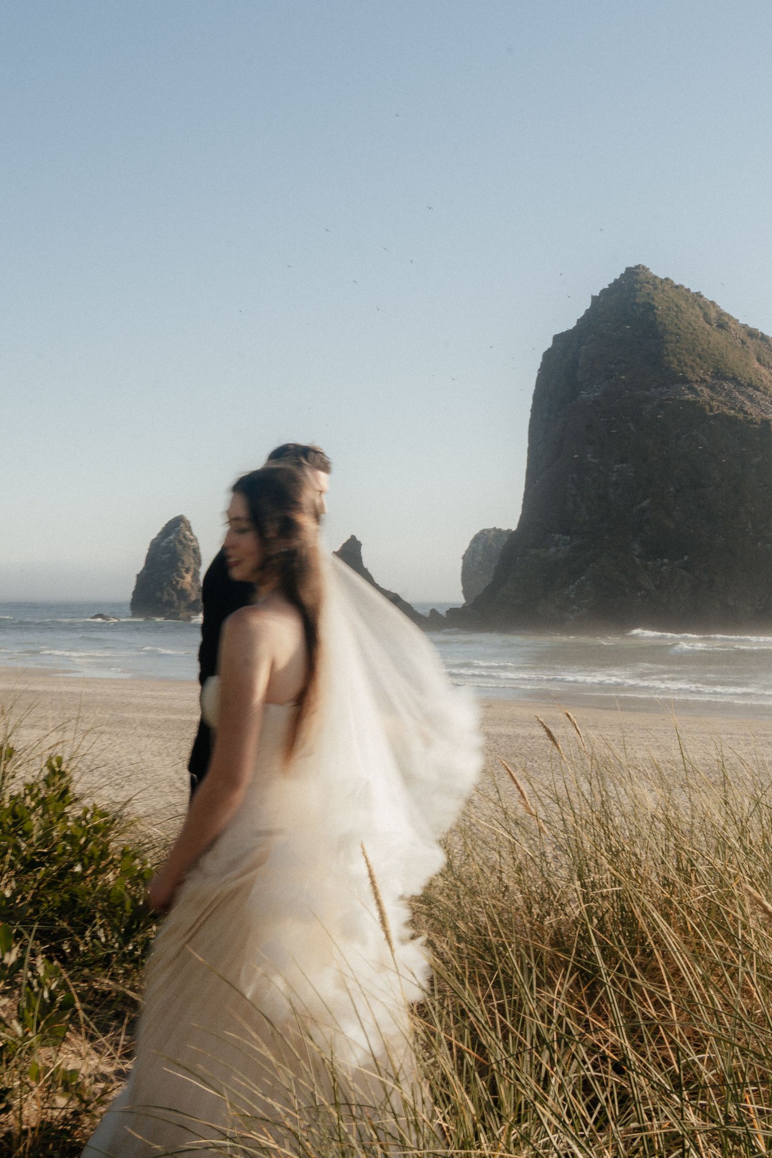 A bride in a flowing white gown and veil stands with her groom on a beach near Haystack Rock in Cannon Beach, Oregon.
