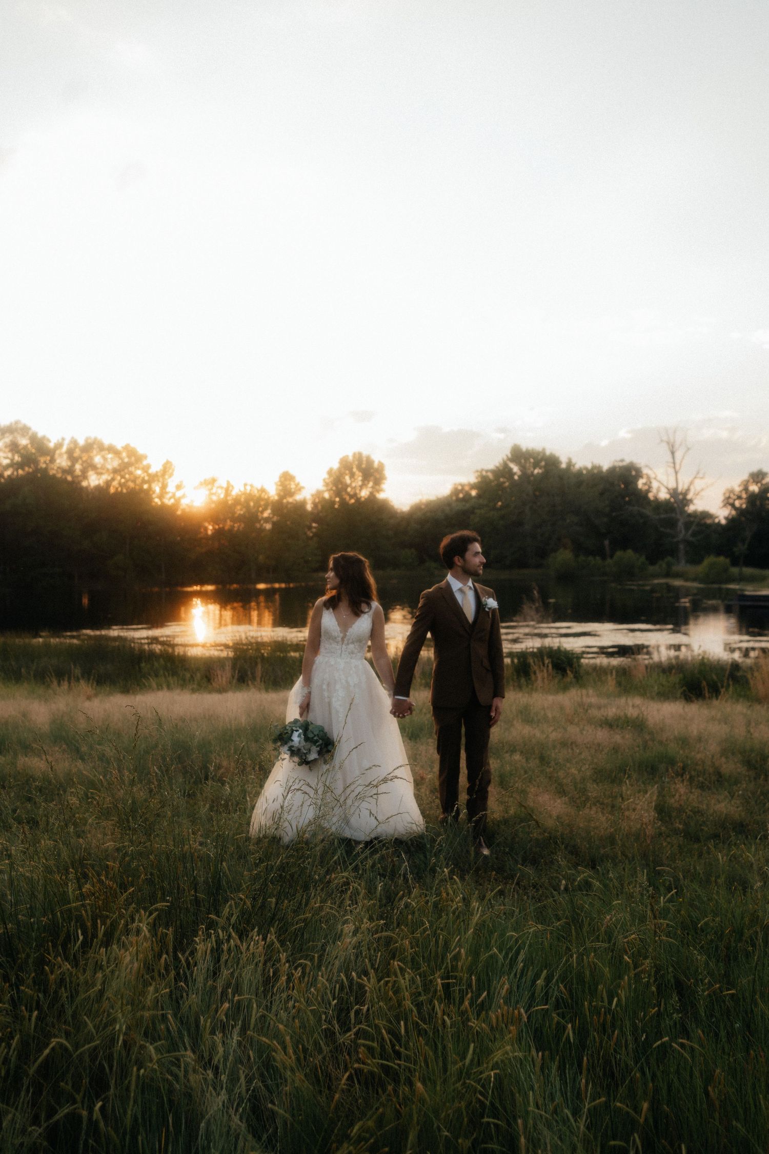 Bride and groom holding hands in a golden sunset field near a lake, bride in white gown with bouquet.