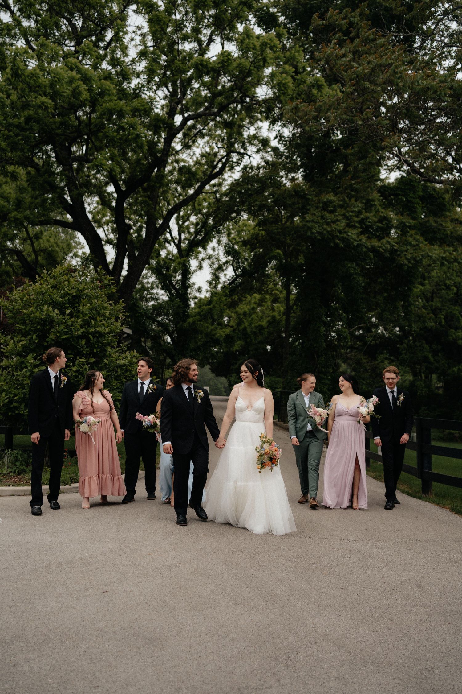 Wedding party walks outdoors on a path surrounded by lush green trees, bride in white gown with bridesmaids in pink dresses.