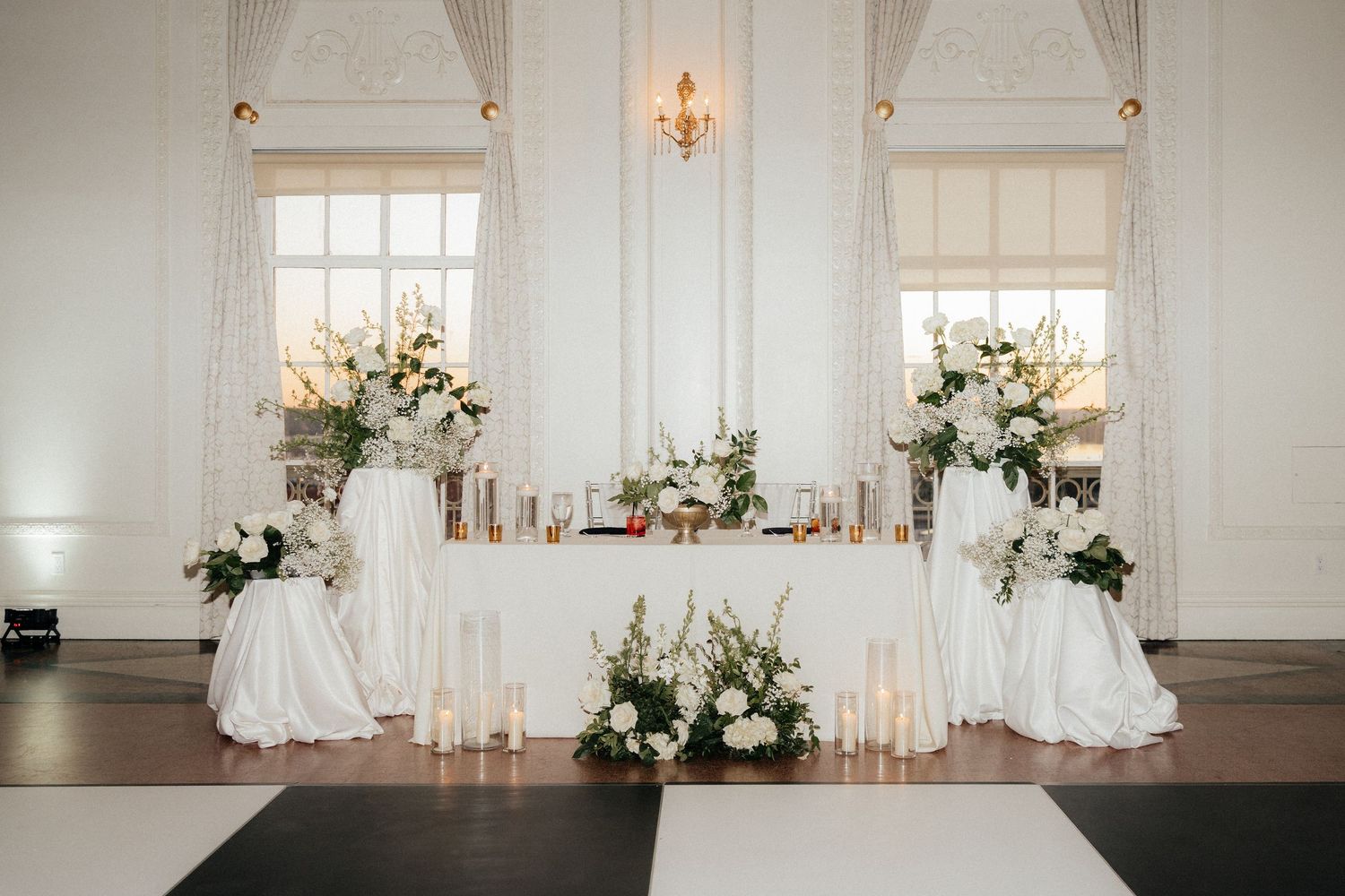 Elegant white wedding ceremony setup with draped curtains, chandeliers, and floral arrangements in a bright ballroom.