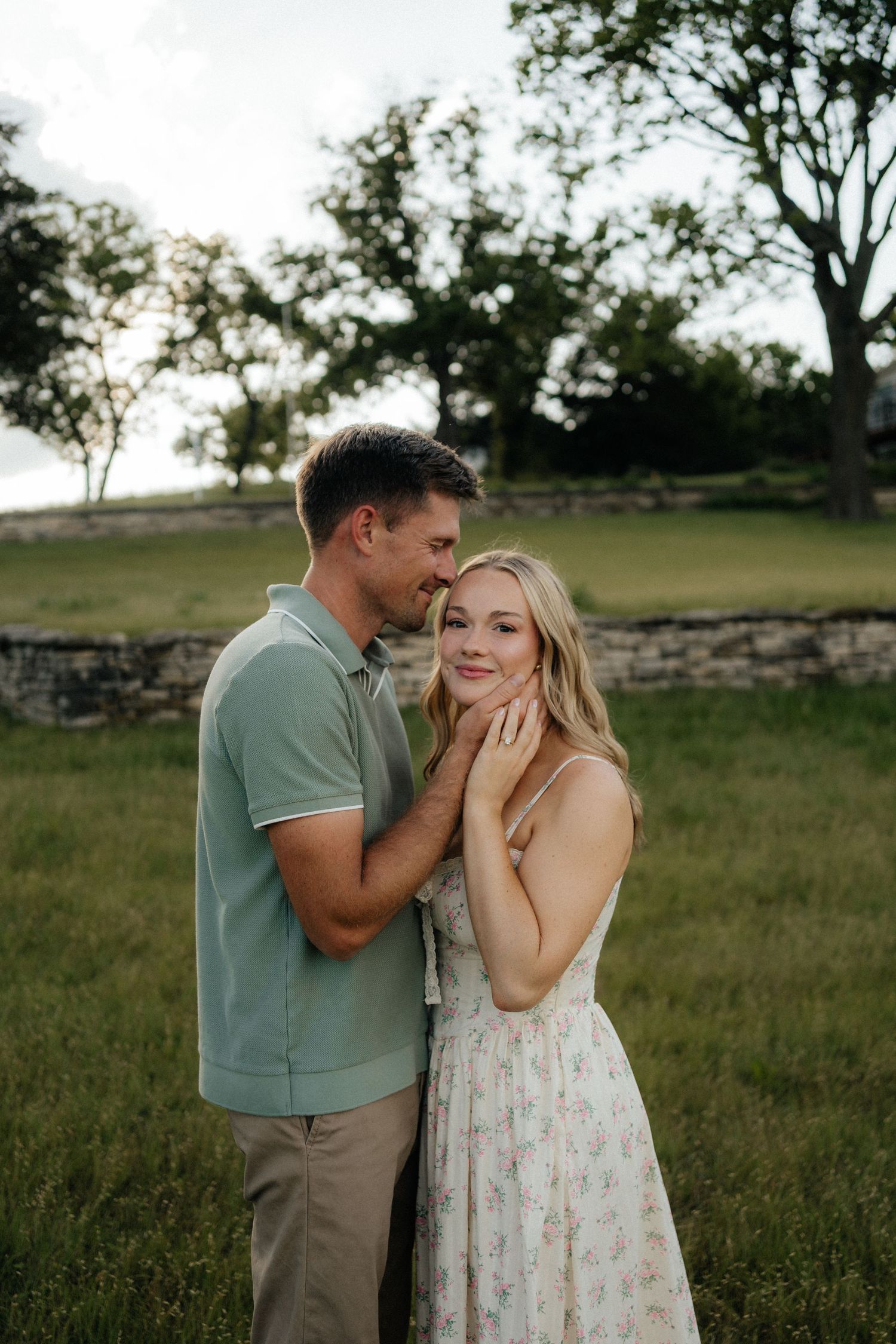 A couple shares a tender moment outdoors, man in green polo kissing woman's temple in a lush green field.