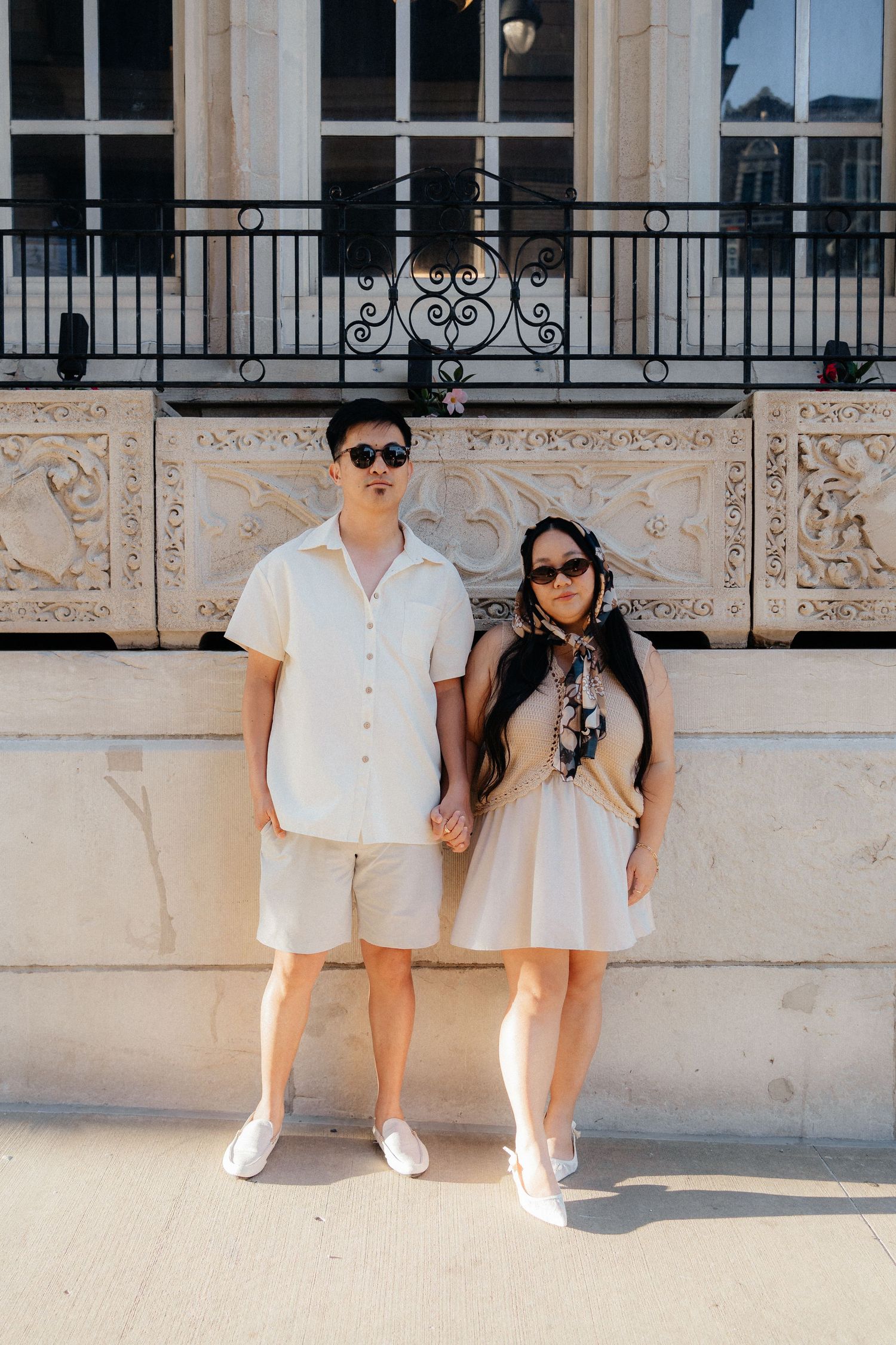 A stylish couple in matching beige outfits and sunglasses hold hands in front of an ornate building facade.