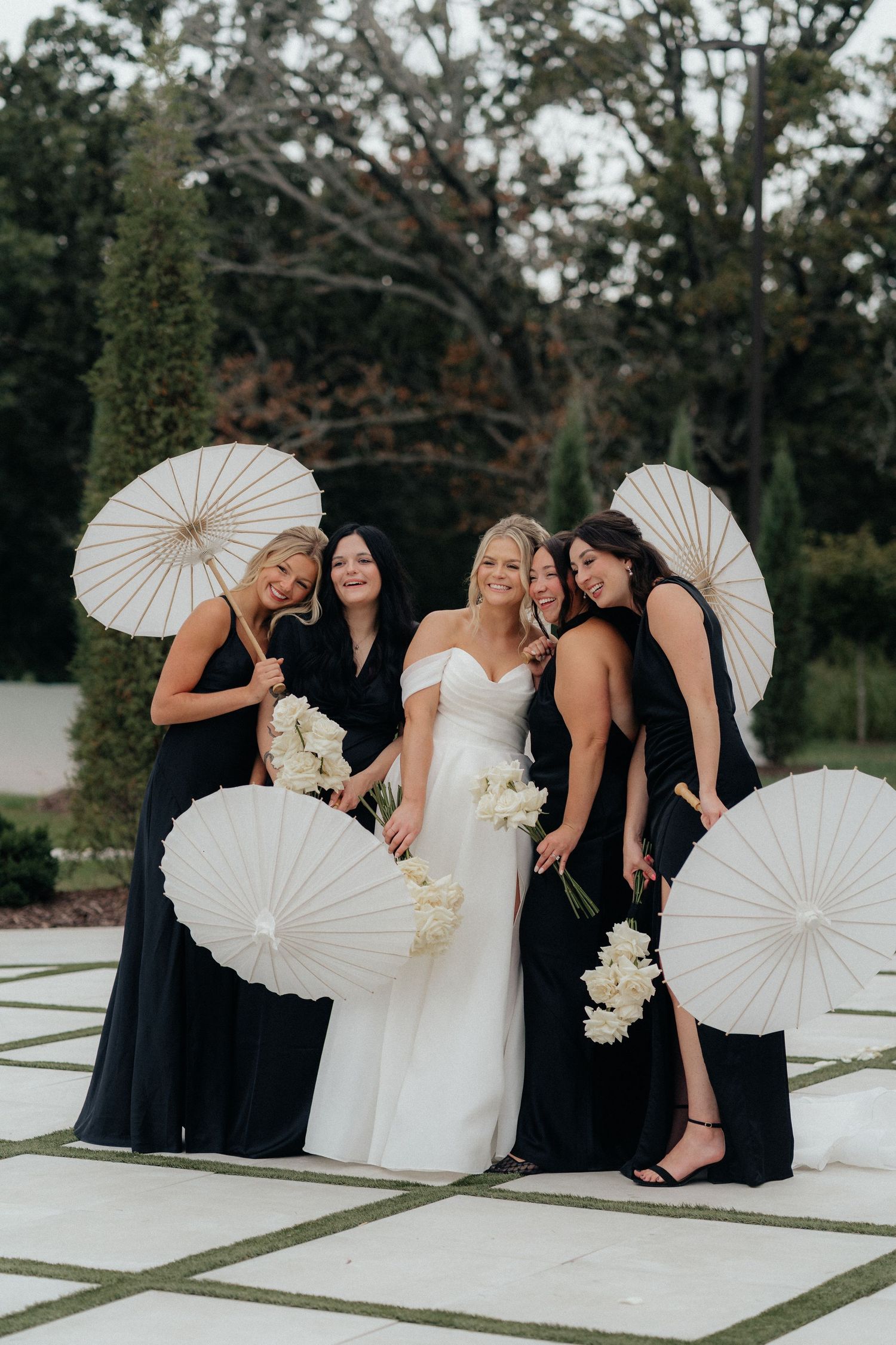 A bridal party in black dresses and white gowns pose together with decorative white paper parasols outdoors.