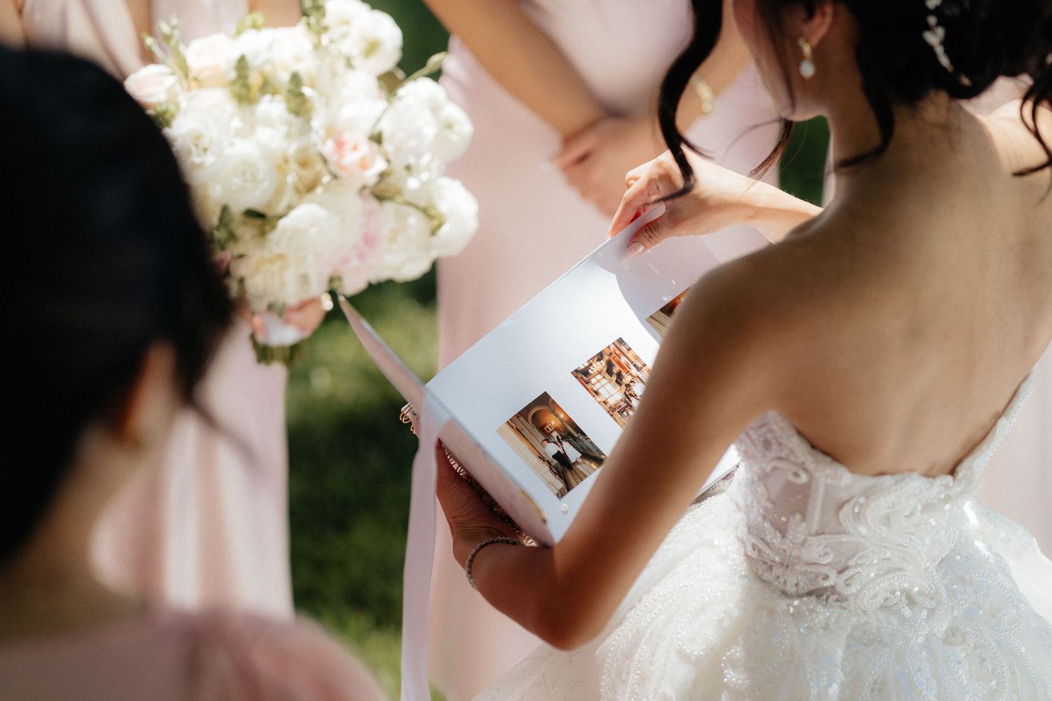 Bride in lace wedding dress holding a photo album with bridesmaid holding pink floral bouquet outdoors.