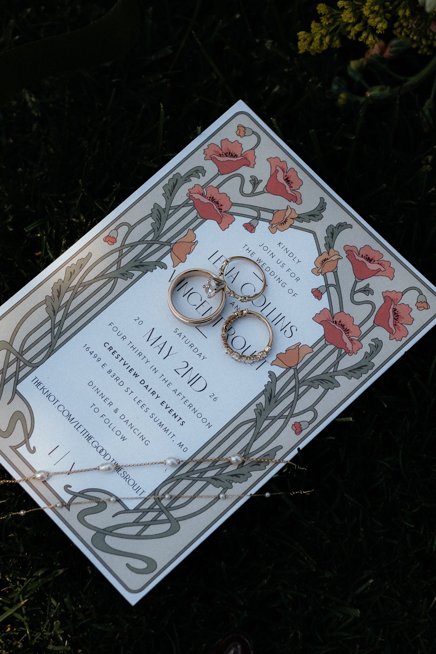 Art nouveau wedding invitation with floral poppy border and two gold wedding rings resting on top, placed on green grass.