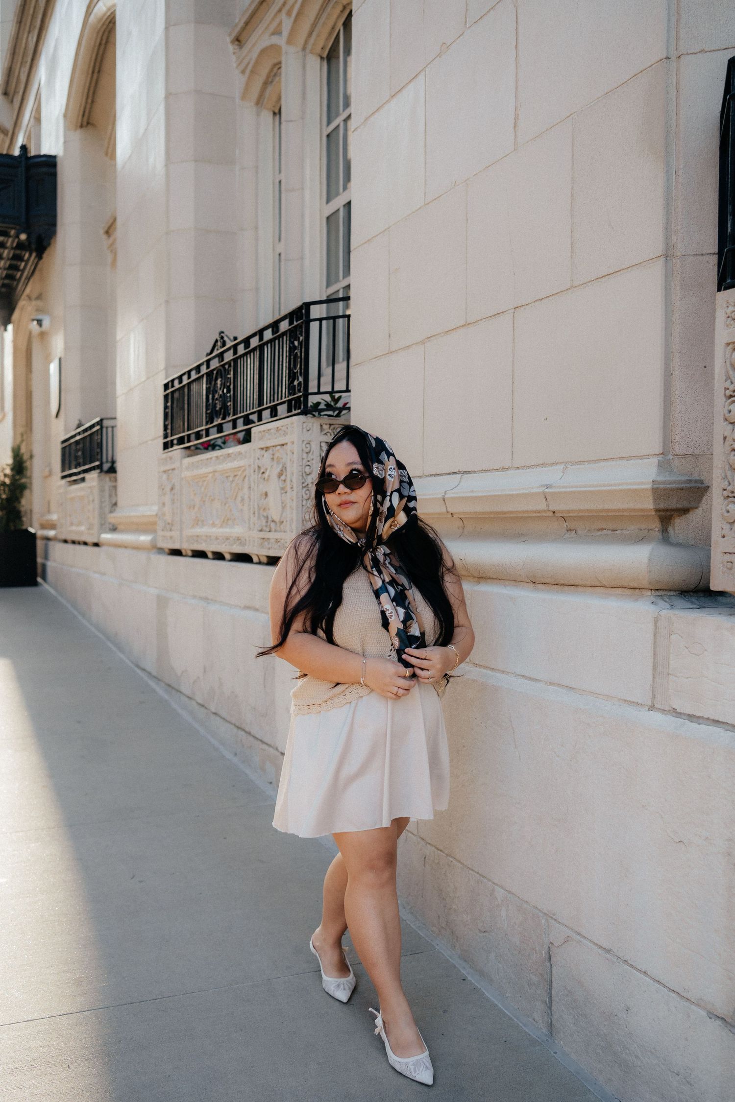 Woman in cream mini dress and heels with floral scarf and sunglasses posing against elegant stone building exterior.