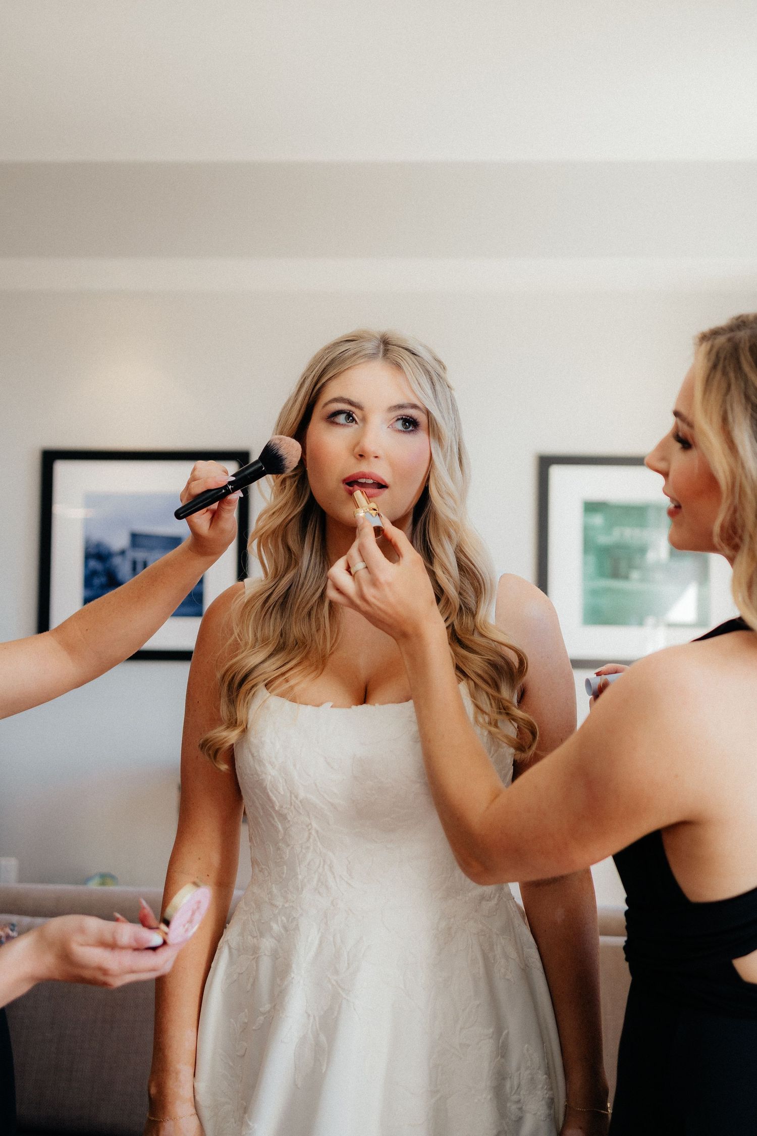 Bride getting final makeup touches applied by beauty team during wedding preparations in white gown.