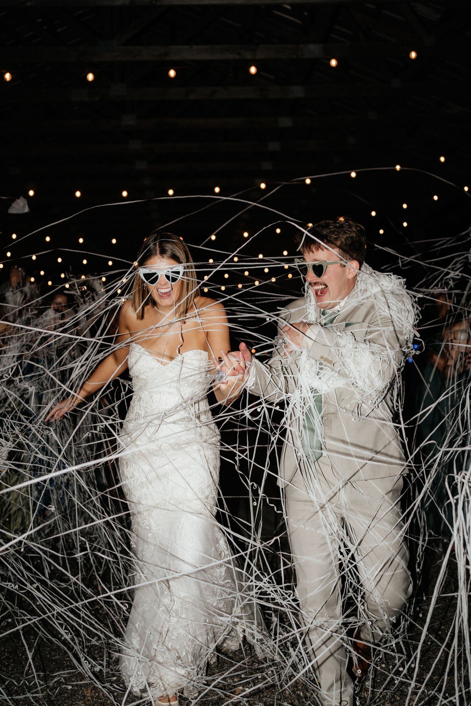 A couple in wedding attire laughs while surrounded by string lights and streamers at a nighttime celebration.