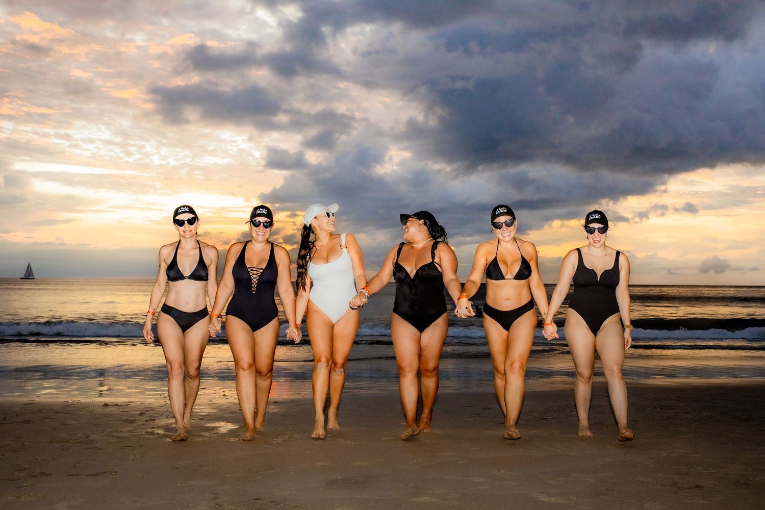 Six women in swimsuits hold hands on a beach at sunset under dramatic cloudy skies. Guanacaste, Costa Rica
