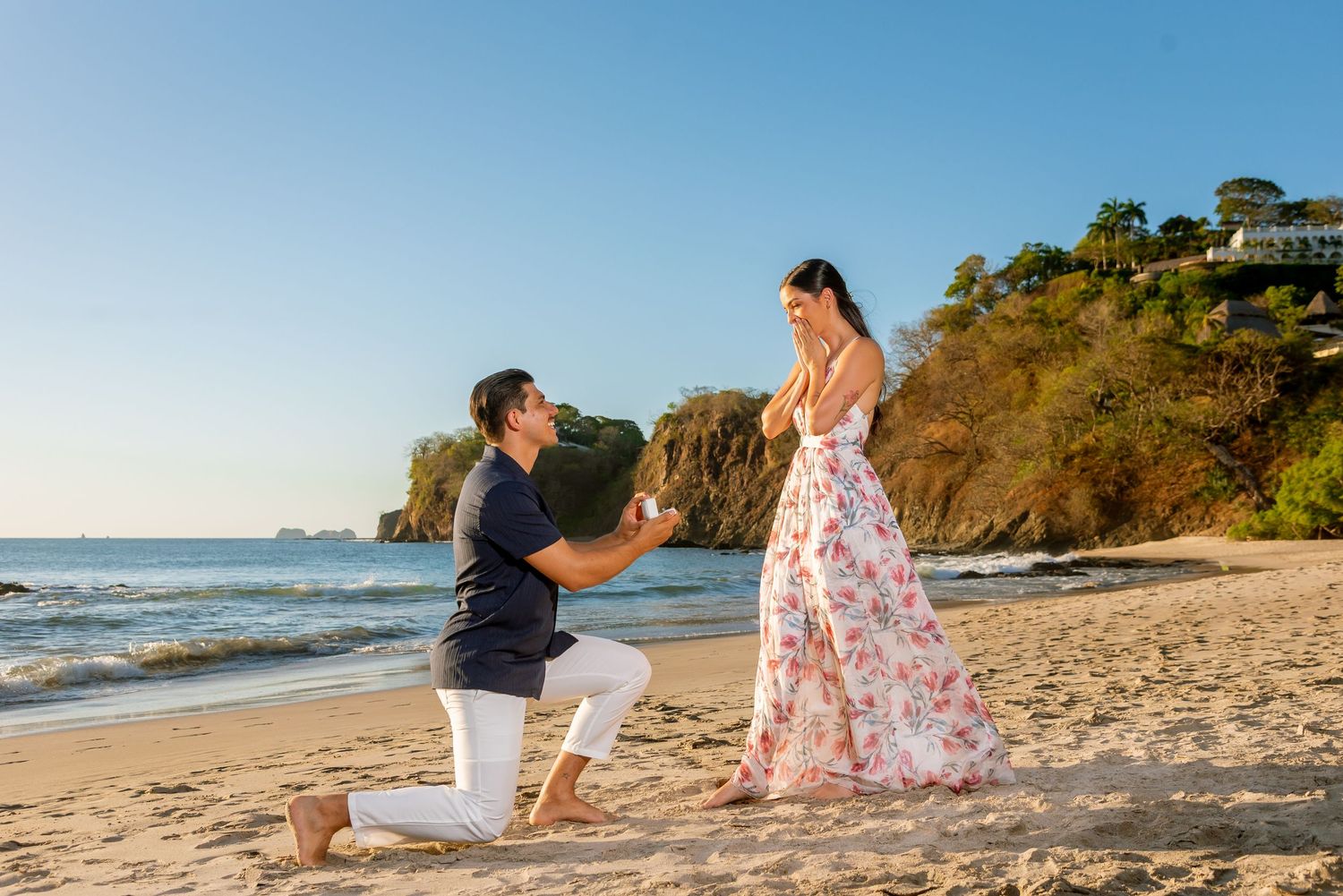 Man proposes on one knee to woman in floral dress on a sunny beach with rocky cliffs in background. Guanacaste, Costa Rica