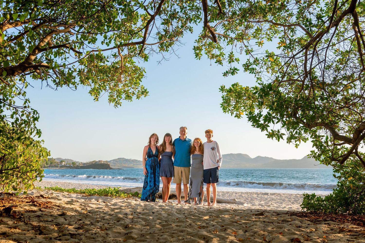 Family of five posing on a tropical beach framed by lush green trees with ocean and mountains in background.