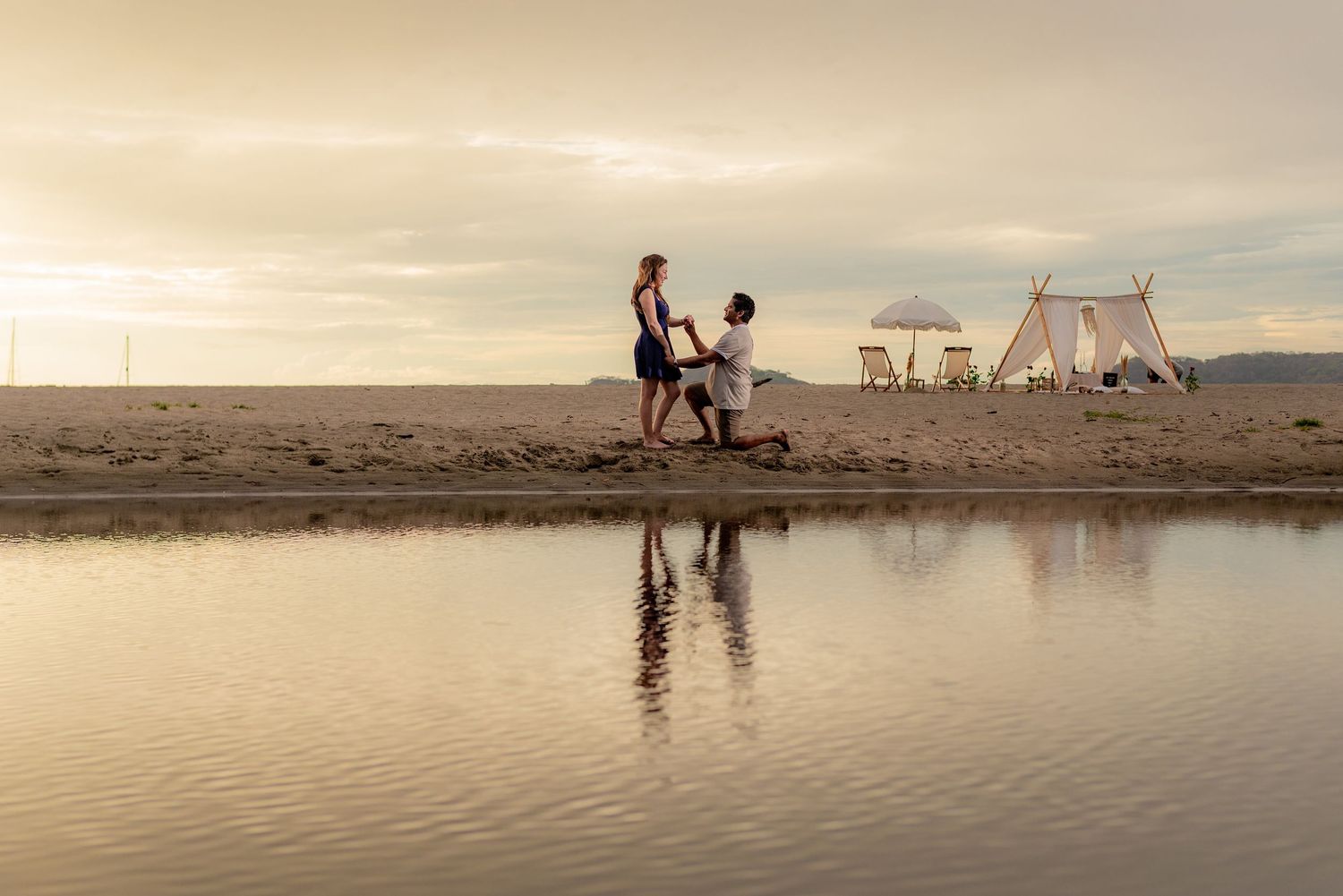 Couple shares a romantic beach proposal at sunset with a decorated canopy setup reflected in calm water. Guanacaste, Costa Rica