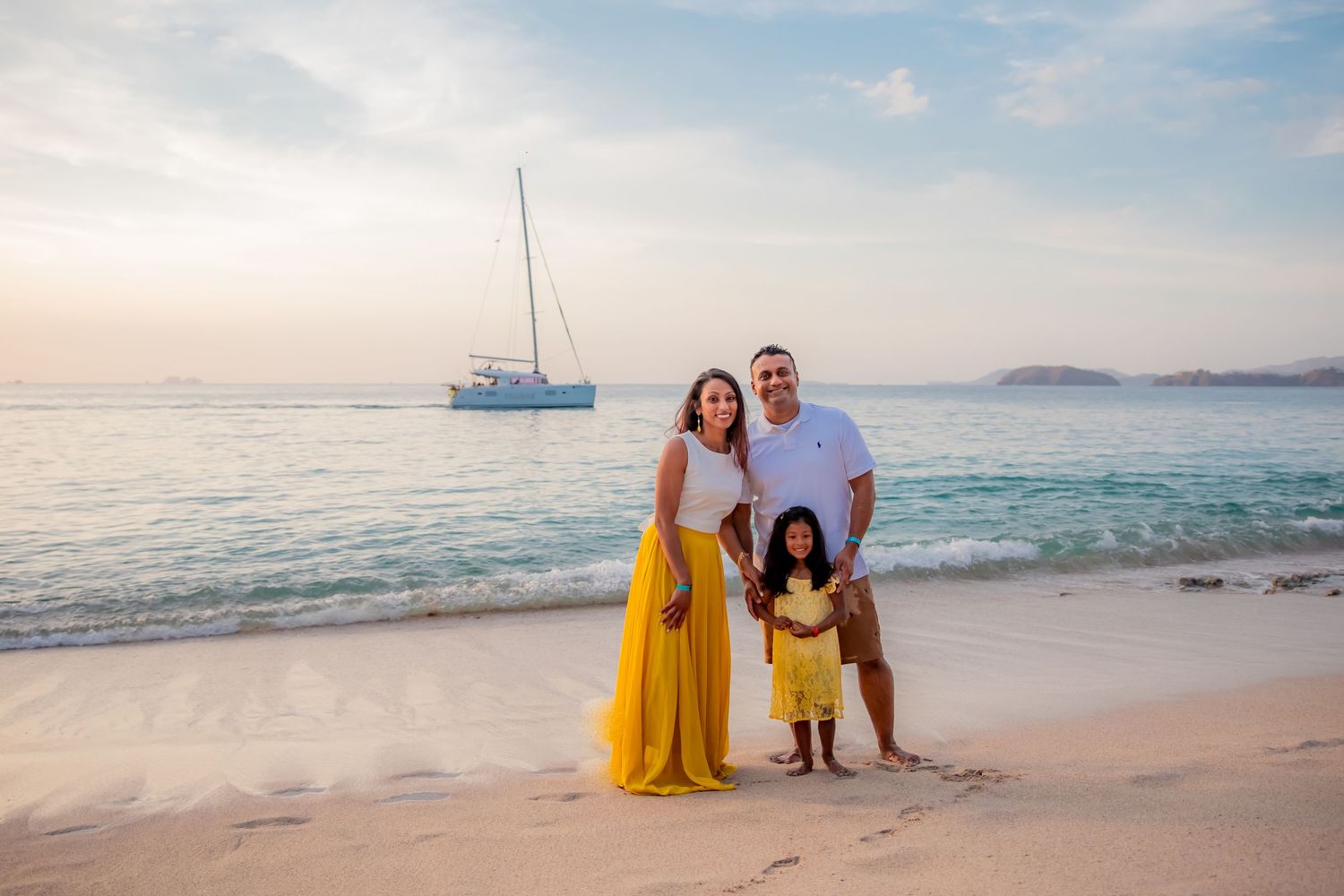 Family of three in yellow outfits posing on white sand beach with sailboat at sunset. Guanacaste, Costa Rica