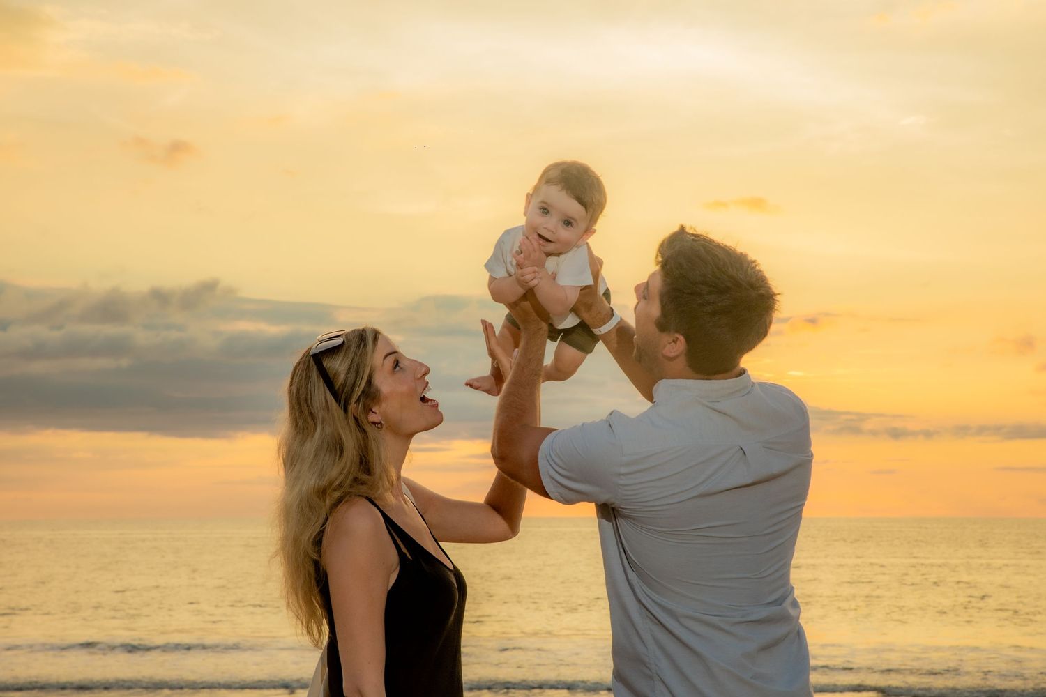 Parents lifting baby at beach during vibrant golden sunset.