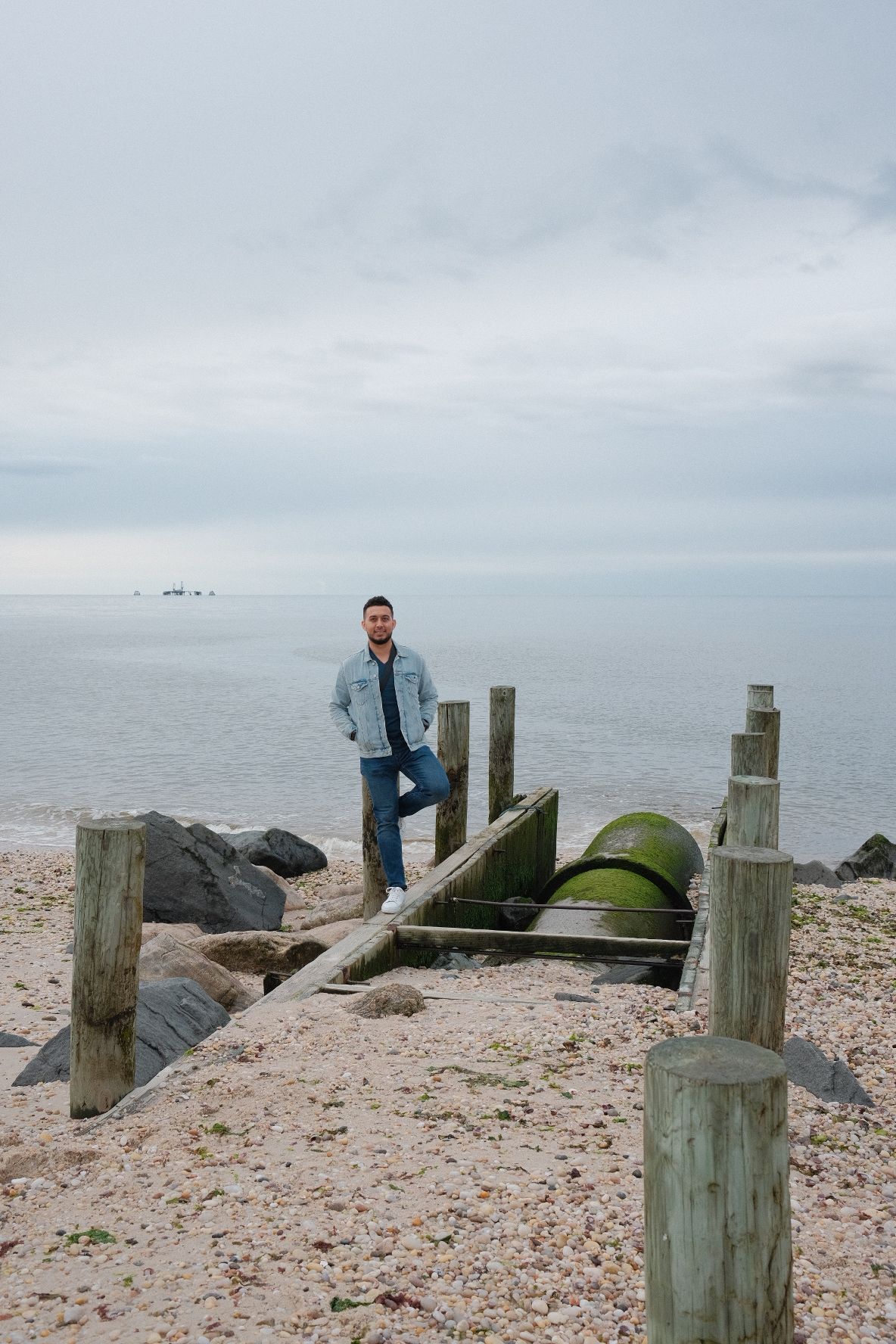 Weathered wooden posts stand along a rocky beach shoreline under an overcast sky.