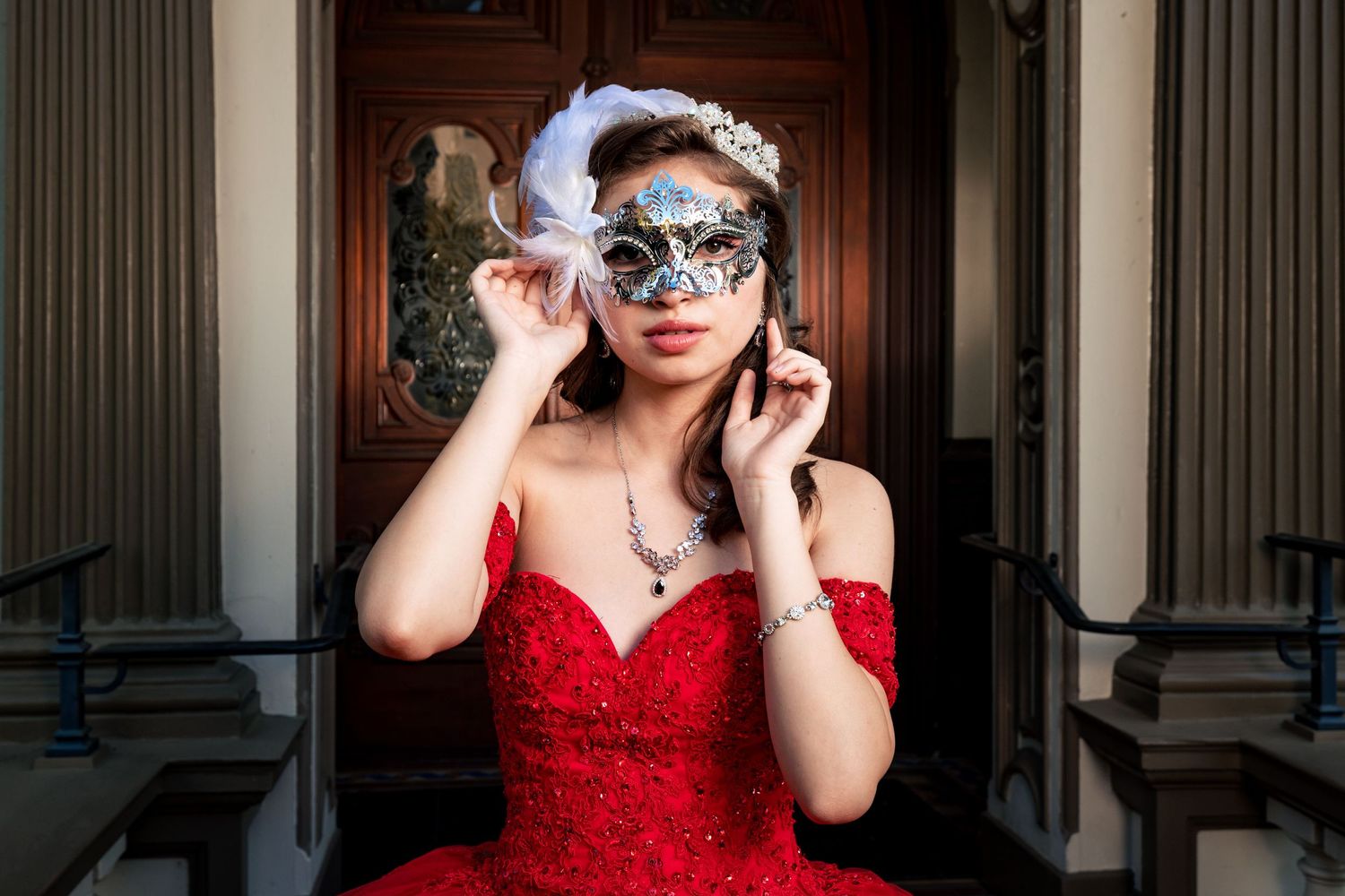 Quinceanera in an elegant red gown and ornate masquerade mask poses in an ornate architectural setting.