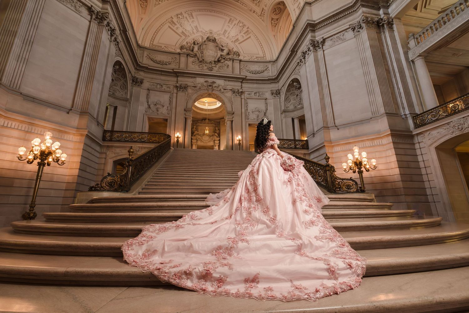 Quinceanera with a dramatic train flows down grand marble stairs in an ornate building with chandelier lighting.