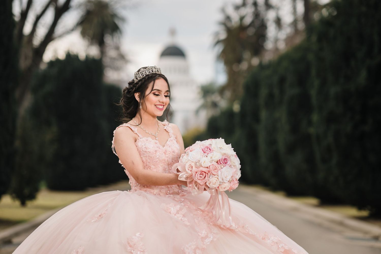 Quinceanera in a pink dress holds a matching pink bouquet in a garden setting at dusk.