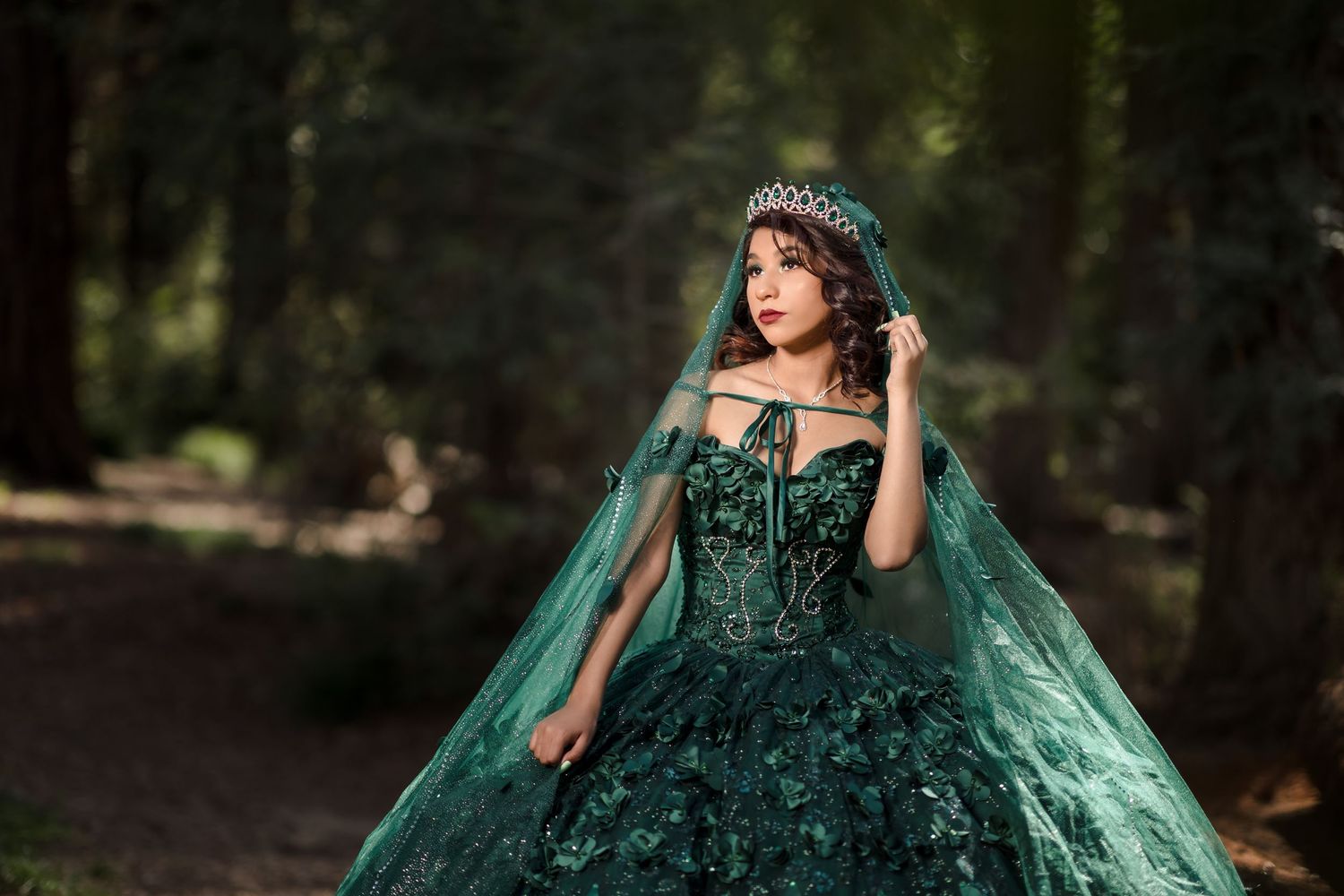Quinceanera wearing an emerald green ball gown with corset top and matching veil poses elegantly in a forest setting.