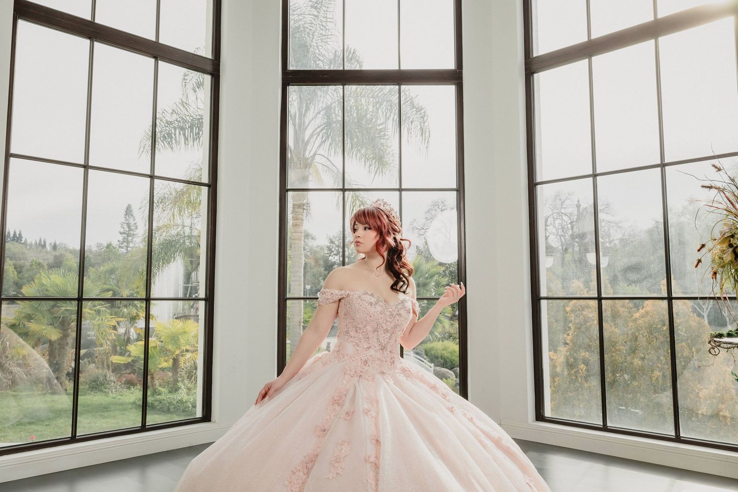 Quinceanera in a blush pink ball gown stands in an elegant sunroom with floor-to-ceiling windows and natural lighting.
