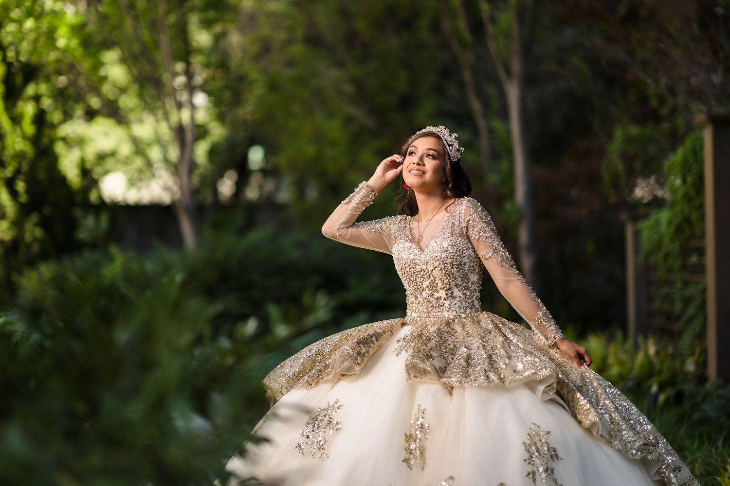 Elegant shimmering quinceanera with long sleeves and floral embellishments photographed in a lush garden setting.