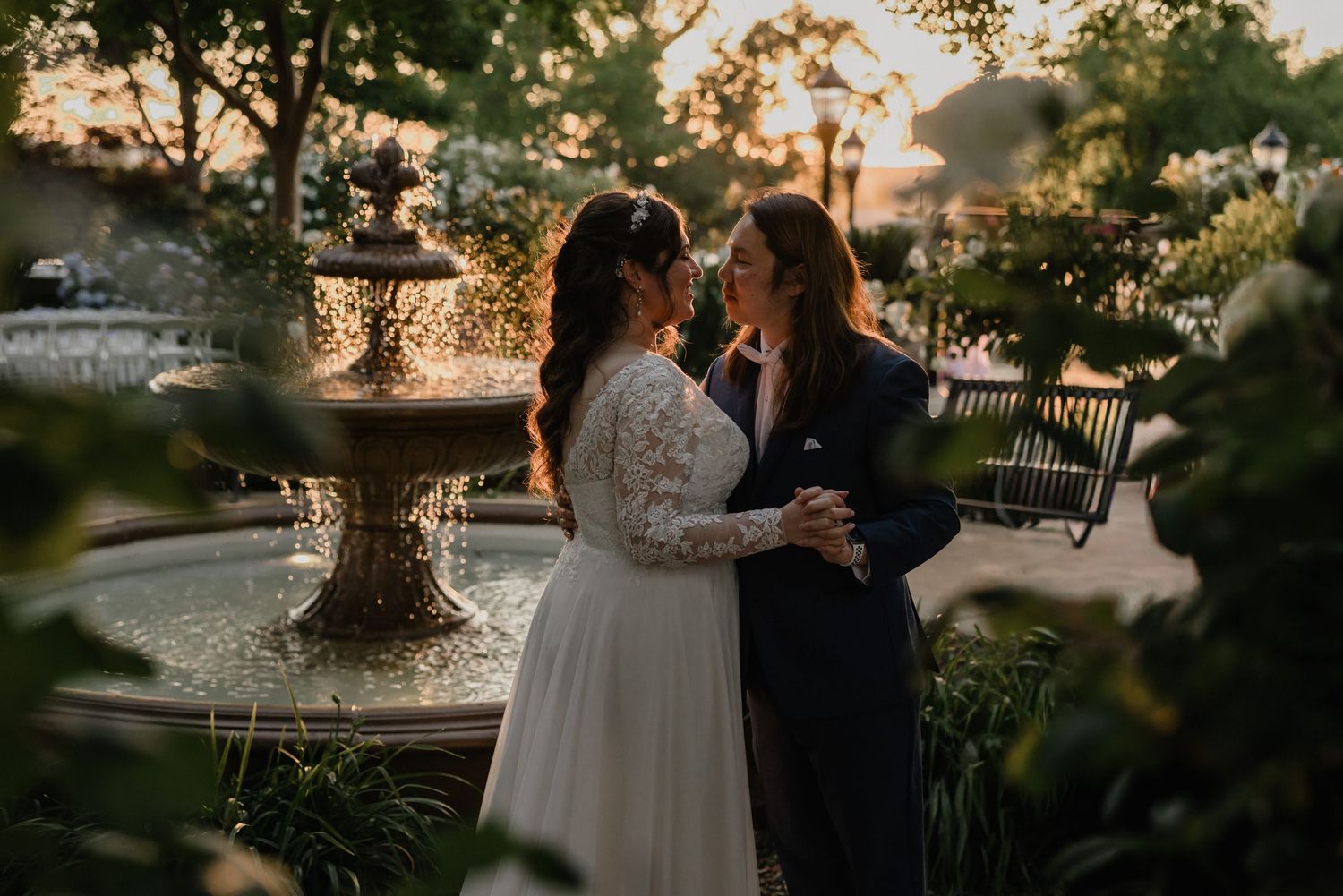 Two brides share a romantic moment by a garden fountain at golden hour, surrounded by lush greenery.