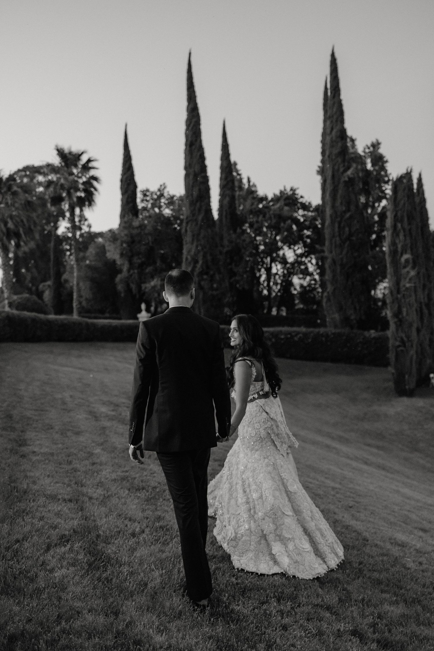 A black and white wedding photo of a couple walking through a garden with tall cypress trees in the background.