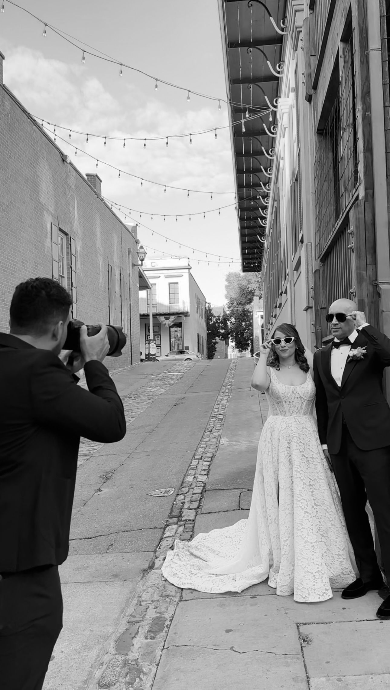 A photographer captures a glamorous wedding moment on a charming cobblestone street with strings of lights overhead.