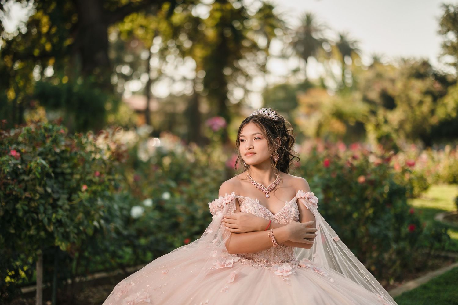 Quinceañera wearing a blush pink off-shoulder gown sits gracefully in a lush garden setting at sunset.