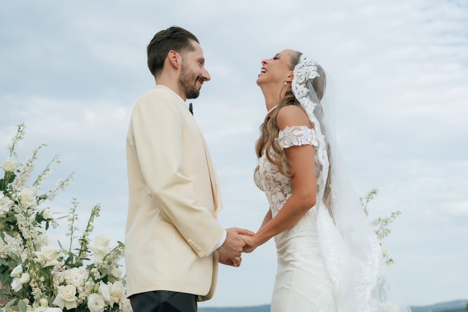 Newlywed couple shares a joyful moment at their outdoor wedding ceremony with white floral arrangements.